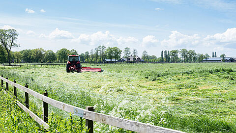 Met een tractor wordt gras gemaaid in een veld. Op de voorgrond een houten hek. 